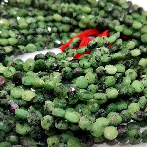 May include: A strand of green gemstone beads with black and red inclusions. The beads are oval shaped and have a faceted finish. The strand is tied with a red cord.