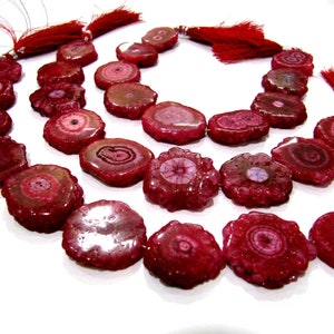 Puede incluir: Una hebra de cuentas de piedra de ágata rosa con una forma natural irregular. Las cuentas tienen un patrón único en capas con un centro blanco y una capa exterior rosa. Las cuentas están enhebradas en un cordón con borlas rojas en cada extremo.