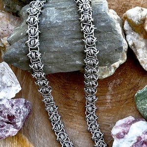 May include: A silver-toned chain necklace constructed from interlocking metal rings in a complex, repeating weave pattern. The chain features a uniform, textured design with a metallic finish, showcasing a series of connected circular links.