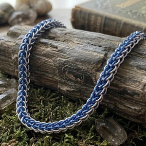 May include: A blue and silver chainmail necklace. The necklace is made of interwoven metal rings, creating a textured, braided appearance. The necklace is displayed on a piece of weathered wood, with moss and crystals in the background.