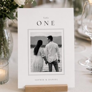May include: A white rectangular table number card featuring the text Table ONE at the top and SOPHIE & DANIEL at the bottom. The centre displays a black and white photograph of a couple holding hands by a lake, framed by thin black lines.