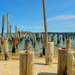 May include: A weathered wooden pier extends into the blue water. The pier is made up of many wooden posts, some of which are broken or missing. The water is calm and clear, and the sky is a bright blue.