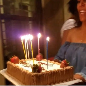 Puede incluir: Un pastel de cumpleaños rectangular con velas encendidas y fresas. El pastel tiene un glaseado marrón claro y está decorado con un chorrito de chocolate. Las velas son de varios colores y el pastel lo sostiene una persona.