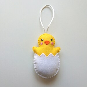 May include: A yellow felt chick ornament with an orange beak and pink cheeks, peeking out of a white felt eggshell. The chick has a white string loop for hanging.
