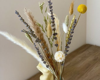 Op de afbeelding: Een doorzichtige glazen fles met een boeket droogbloemen, waaronder paarse lavendel, beige pampasgras, groene tarwestengels, een gele craspedia en witte pluizige konijnenstaartjes, met daarnaast een crèmekleurige kaars in de vorm van een torso.