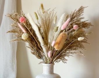 Può includere: Un bouquet di erba della pampa essiccata, piume bianche soffici e palline rosa disposto in un vaso di ceramica color crema. Una capsula di cotone bianco e una piccola candela bianca a forma di cubo a bolle si trovano accanto al vaso.