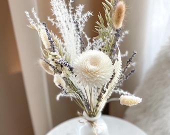 Op de afbeelding: Een boeket droogbloemen in een witte keramische fles. Het arrangement bevat een centrale witte chrysant, takjes paarse lavendel, pluizig wit pampasgras, groene haverstengels en beige konijnenstaartjes, samengebonden met een dun touwtje.