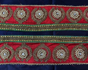 May include: Embroidered trim in red and gold, featuring a repeating pattern of ornate medallions. The trim has a green border with gold accents, and is embellished with pearls and clear stones. The background is a dark blue fabric.