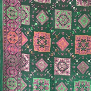 May include: A green fabric with a repeating pattern of pink, orange, and silver geometric shapes. The fabric has a border with a more intricate design.