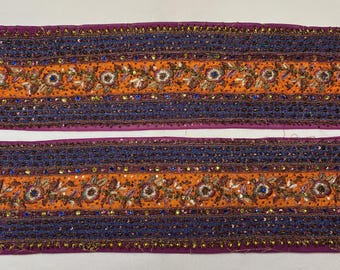 May include: Two strips of decorative fabric with a repeating pattern. The design features alternating bands of orange and dark blue, embellished with floral embroidery and small, colorful beads. The edges are trimmed with a purple border.