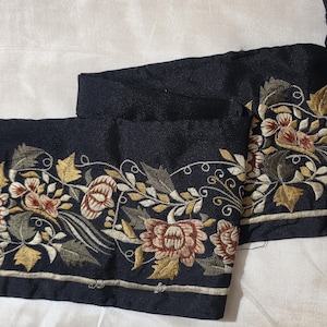 May include: A black fabric strip featuring an intricate embroidered floral border. The design includes stylised flowers and leaves in shades of muted gold, olive green, and soft pink, accented with thin, swirling cream-coloured thread lines.