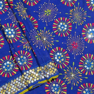 May include: A vibrant blue fabric featuring an embroidered pattern of colourful starbursts in red, yellow and green thread. The design includes circular motifs accented with reflective silver sequins, and a border lined with rows of metallic silver sequins.