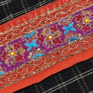 May include: A close-up of a colorful embroidered trim with sequins and beads. The trim features a purple base with red and gold floral designs, and a red base with gold and orange floral designs.