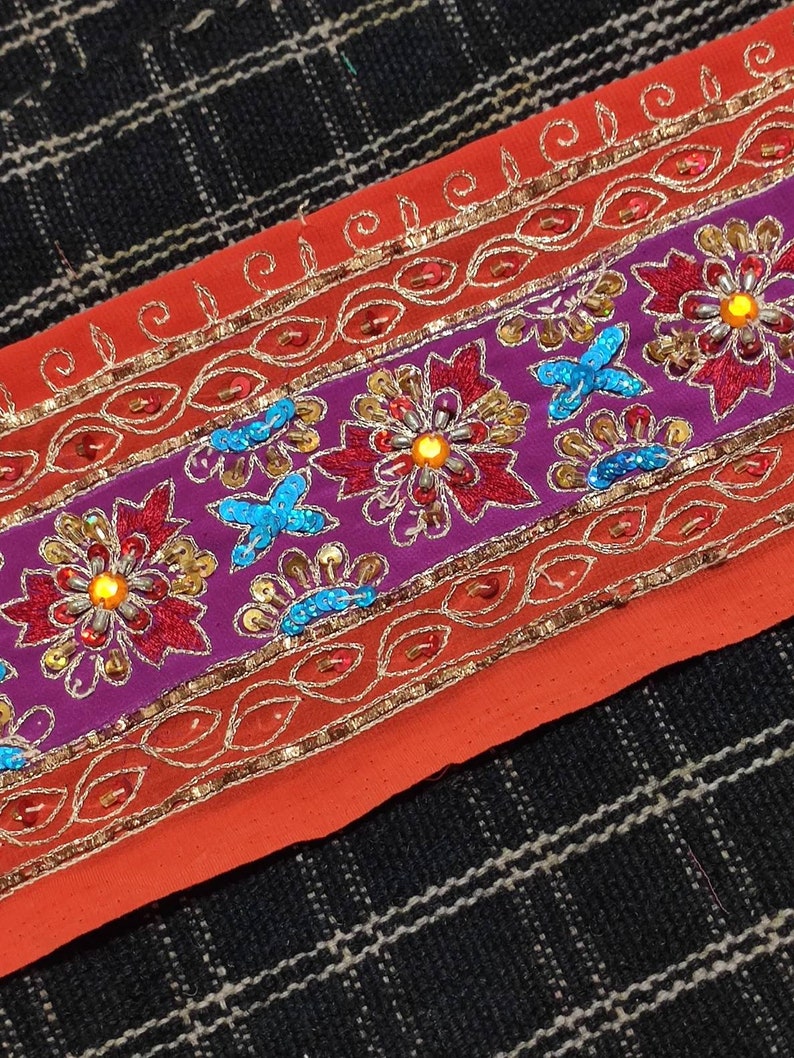 May include: A close-up of a colorful embroidered trim with sequins and beads. The trim features a purple base with red and gold floral designs, and a red base with gold and orange floral designs.