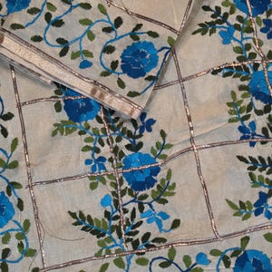 Könnte beinhalten: Ein cremeweißer Stoff mit einem Gittermuster aus metallisch goldfarbenen Bandstreifen. Der Stoff ist mit gestickten Blumenmotiven in leuchtendem Blau und Dunkelgrün verziert, die blühende Blumen und blättrige Ranken darstellen.
