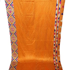 Op de afbeelding: Een rechthoekig oranje textiel met een centraal ruitpatroon. De randen zijn afgewerkt met ingewikkeld geometrisch borduurwerk in levendige tinten roze, blauw, wit en geel, met een bruine bies aan de boven- en onderkant.