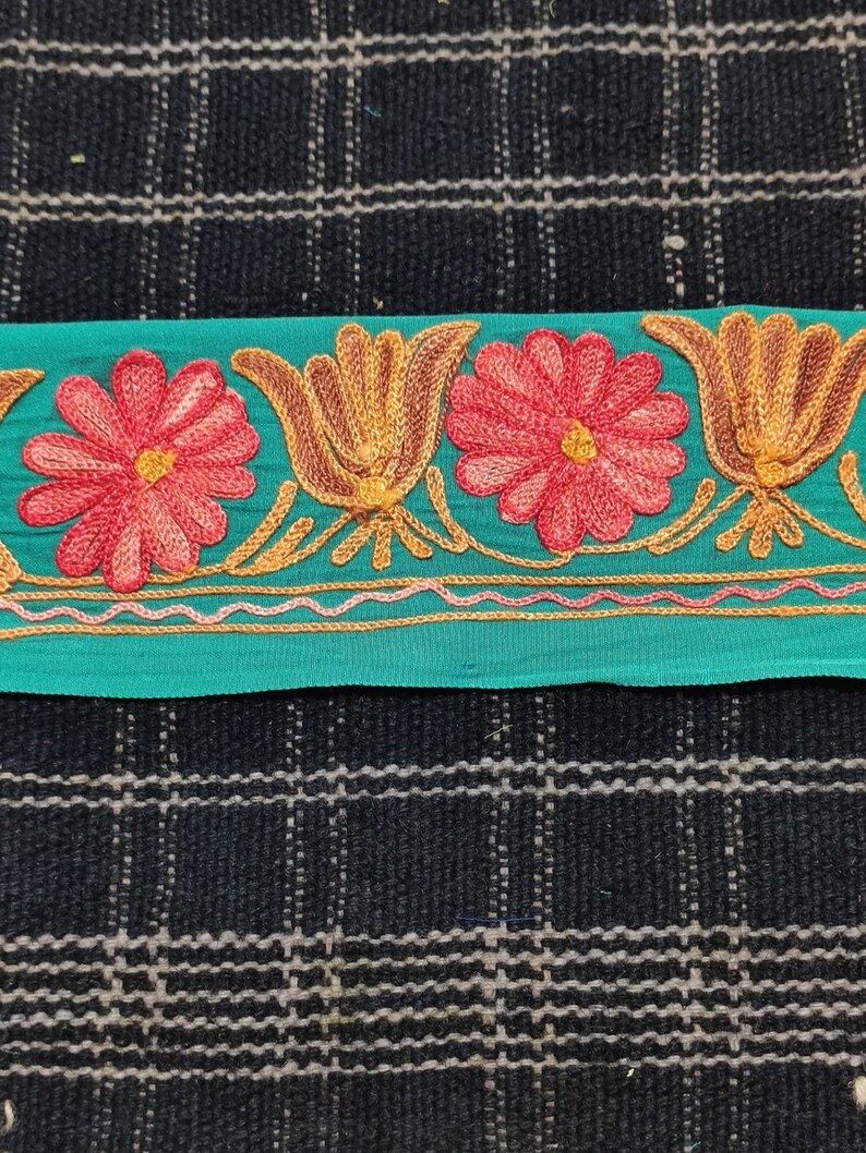 May include: A teal blue fabric trim with embroidered red flowers and gold leaves. The trim has a scalloped edge and a gold thread design.