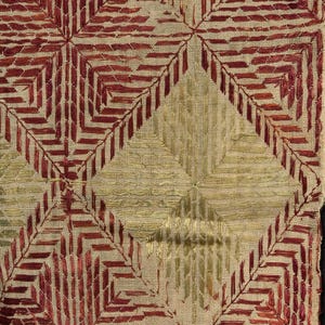 Könnte beinhalten: Ein gewebter Stoff mit einem sich wiederholenden geometrischen Muster aus Rauten und Quadraten. Das Design besteht aus dunkelrotem und goldfarbenem Stickgarn, das auf ein naturbeiges, grob strukturiertes leinenartiges Grundmaterial gestickt ist.