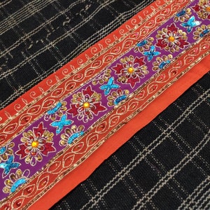 May include: A close-up of a colorful embroidered trim with red, orange, purple, and blue accents. The trim features floral and butterfly designs with sequins and beads.