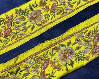 May include: Two yellow fabric trims with floral embroidery and beaded accents. The trim features a repeating pattern of flowers and leaves in shades of red, green, and gold.