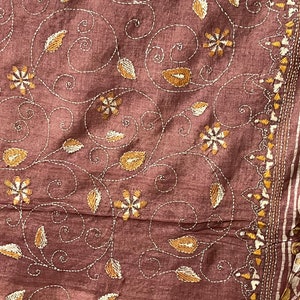 May include: A brown fabric with a floral embroidery pattern in white and gold thread. The design features swirling vines, leaves, and flowers.