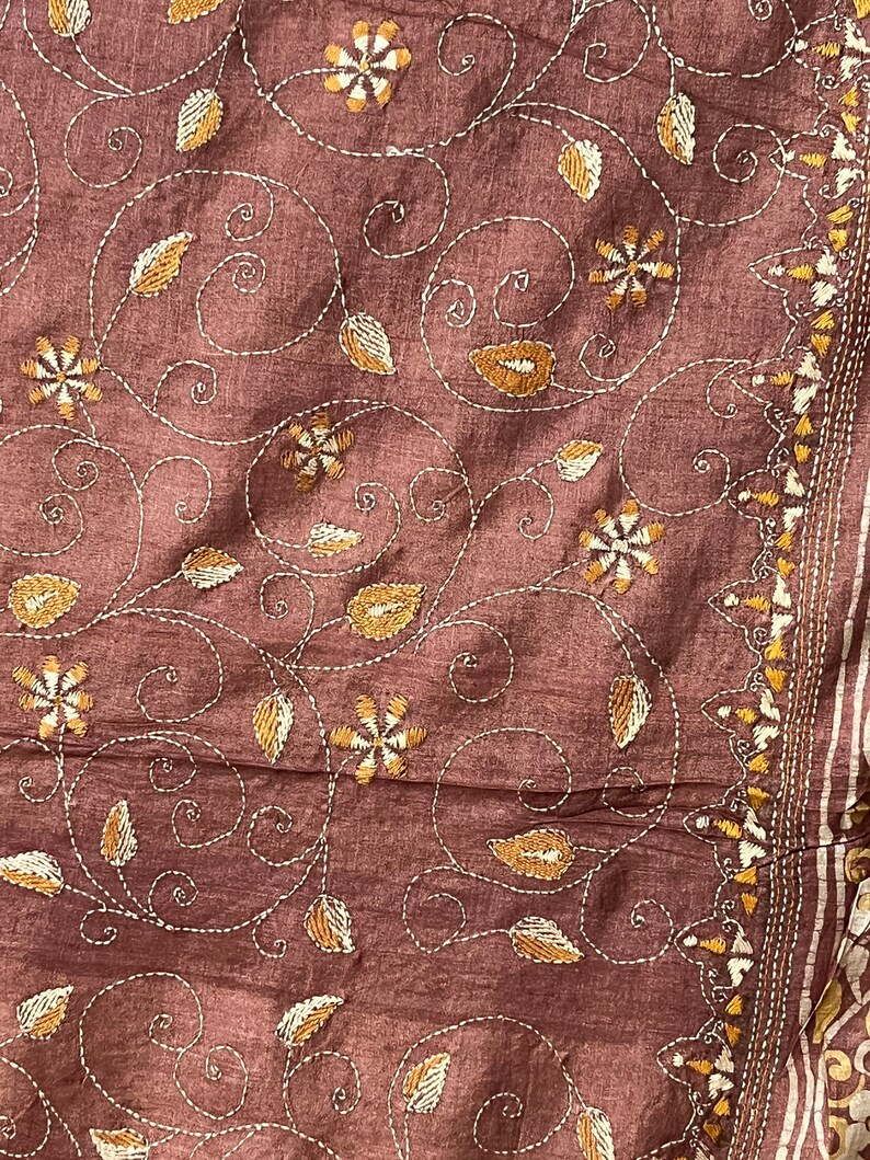 May include: A brown fabric with a floral embroidery pattern in white and gold thread. The design features swirling vines, leaves, and flowers.