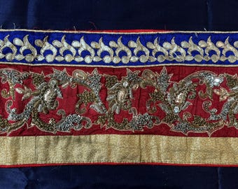 May include: Close-up of a decorative textile trim. The trim features a blue border with a white floral design, a red central section with ornate gold and silver embroidery, and a gold lower border. The background is dark blue.