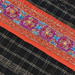 May include: A close-up of a colorful embroidered trim with sequins and beads. The trim is on a red fabric background and features a floral design with purple, red, orange, and blue accents.