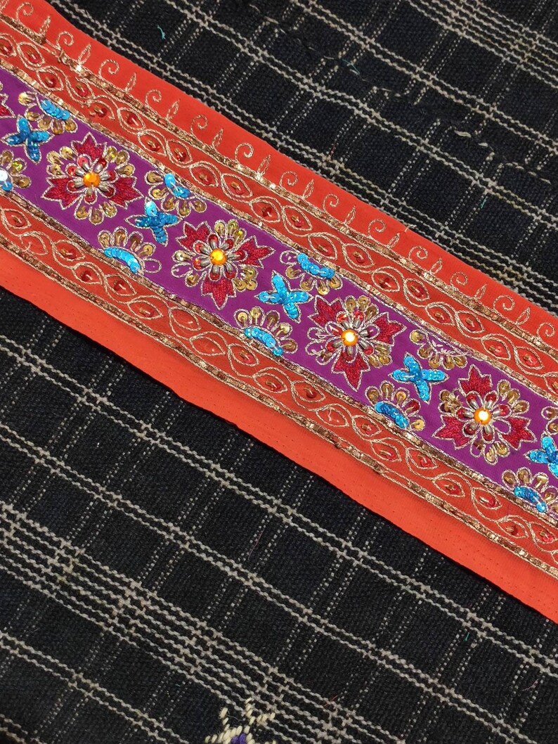 May include: A close-up of a colorful embroidered trim with sequins and beads. The trim is on a red fabric background and features a floral design with purple, red, orange, and blue accents.