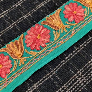 May include: Teal fabric trim with embroidered flowers in shades of pink and orange. The trim features a repeating pattern of stylised flowers and leaves.