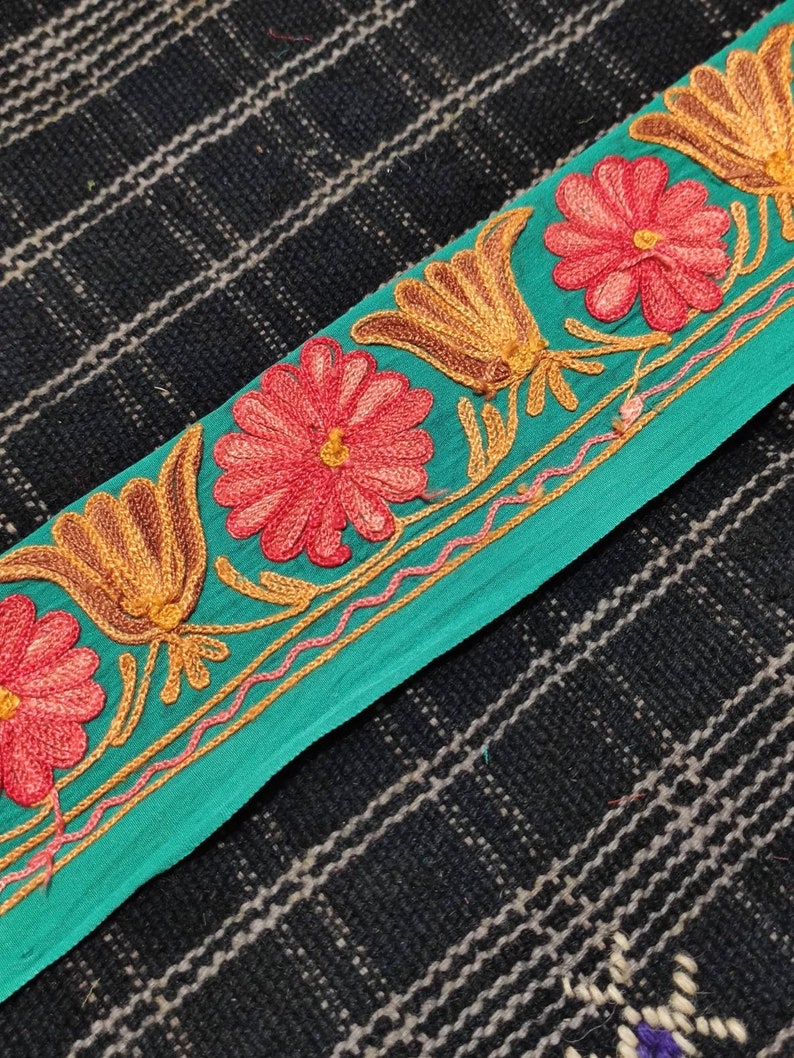 May include: Teal fabric trim with embroidered flowers in shades of pink and orange. The trim features a repeating pattern of stylised flowers and leaves.