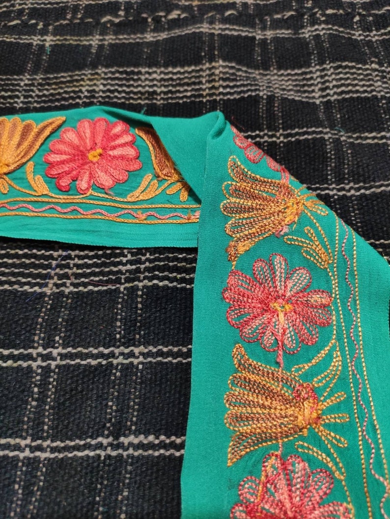 May include: Teal fabric trim with gold and pink embroidered floral designs. The trim features a repeating pattern of flowers and leaves.