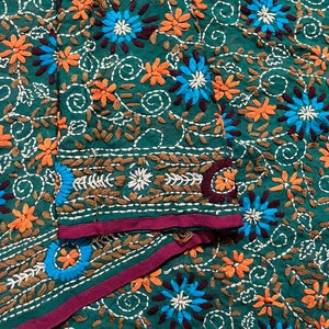 Könnte beinhalten: Nahaufnahme eines grünen Stoffes mit aufwendiger Stickerei. Das Design zeigt orange und blaue Blumen, weiße Nähte und braune Blätter. Ein dunkelvioletter Rand akzentuiert die Stoffkanten und schafft ein farbenfrohes Textil.