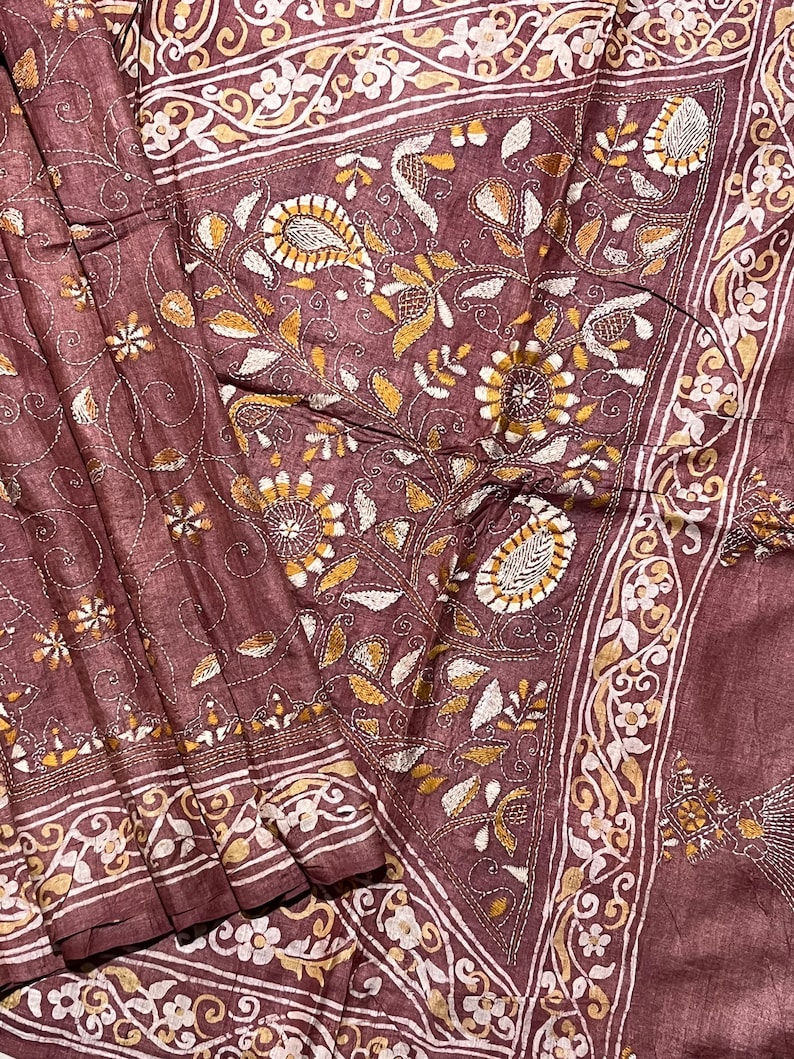 May include: A brown silk saree with intricate white and gold embroidery. The embroidery features floral patterns and paisley designs.