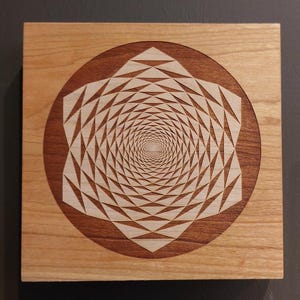 Pode incluir: Obra de arte quadrada em madeira com um design geométrico. A obra apresenta uma base de madeira clara com uma borda circular marrom escuro. Dentro da borda, há um padrão espiral detalhado em um tom de madeira mais claro.