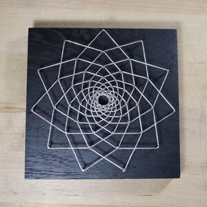 May include: A geometric design created with string wrapped around nails hammered into a black wooden board.