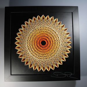 May include: A framed string art piece with a geometric design in shades of orange and yellow. The design is a mandala with a black center and a white background. The frame is black.