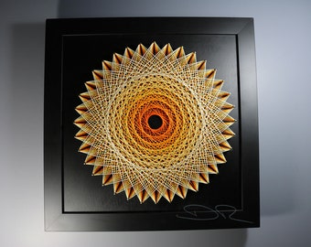 May include: A framed string art piece with a geometric design in shades of orange and yellow. The design is a mandala with a black center and a white background. The frame is black.