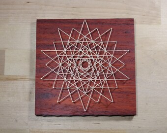 May include: A geometric design created with string art on a dark red wooden board. The design is a starburst pattern with a smaller starburst in the center.