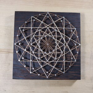 May include: A geometric design created with string art on a dark wood background. The design features a series of intersecting lines and shapes, creating a complex and intricate pattern.