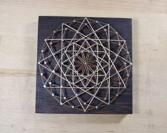 May include: A geometric design created with string art on a dark wood background. The design features a series of intersecting lines and shapes, creating a complex and intricate pattern.