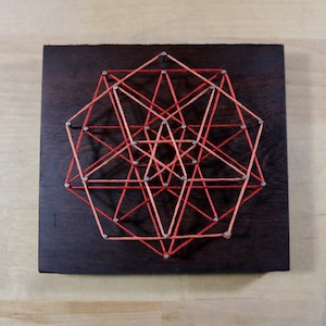 May include: A geometric design created with string art on a dark wood background. The design features a series of triangles and squares connected by red and orange string.