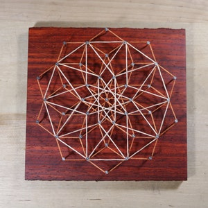 May include: A geometric design created with string and nails on a dark wood background. The string is white and brown, creating a complex pattern.