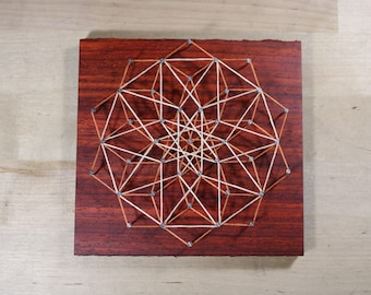 May include: A geometric design created with string and nails on a dark wood background. The string is white and brown, creating a complex pattern.