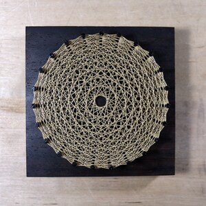 May include: A handmade string art piece featuring a circular design. The artwork is crafted with beige string on a dark stained wooden square. Small black beads are placed around the outer edge of the circle.