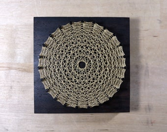 May include: A handmade string art piece featuring a circular design. The artwork is crafted with beige string on a dark stained wooden square. Small black beads are placed around the outer edge of the circle.