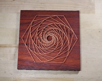 May include: A copper wire string art piece on a dark wood background. The wire is wrapped around nails in a spiral pattern to create a geometric design.