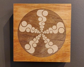 May include: Wooden square with a circular design. The design features a central starburst pattern composed of interconnected, light-colored, abstract shapes set against a darker wood grain background. The square is made of light-colored wood.