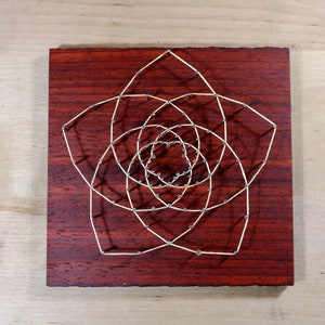 May include: A geometric design created with string wrapped around nails hammered into a dark red wooden board.