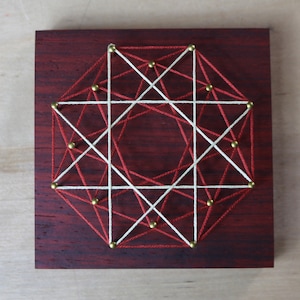 May include: A geometric design created with red and white string wrapped around gold tacks on a dark wood background.